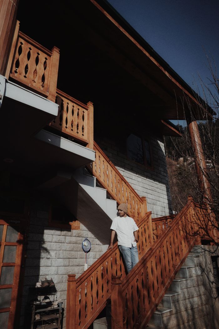 service-02 A man in casual attire stands on the wooden steps of a rustic house, enjoying the daylight.