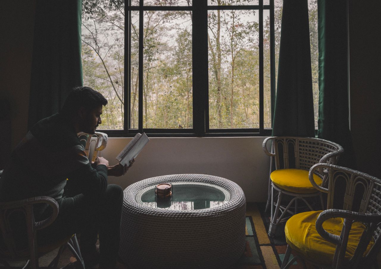 A man reads near a window with wicker chairs and a glass-topped table.