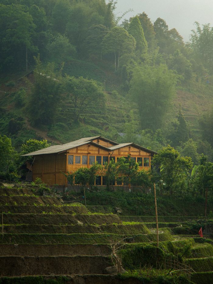service-04 A rustic wooden house surrounded by terraced fields and lush green hills in a serene landscape.